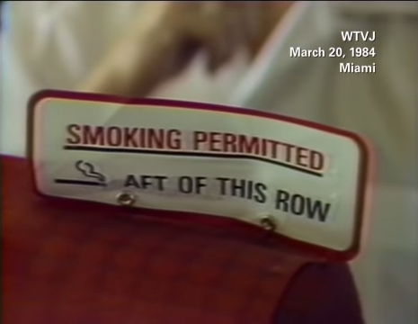 Sign on airplane seat reading "SMOKING PERMITTED AFT OF THIS ROW" with date March 20, 1984, from WTVJ Miami