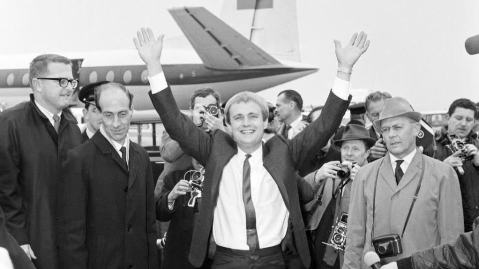 david mccallum with both arms raised to greet fans in front of an airplane