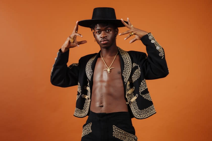 A man in a black cowboy hat and gold-accented black jacket posing against an orange background