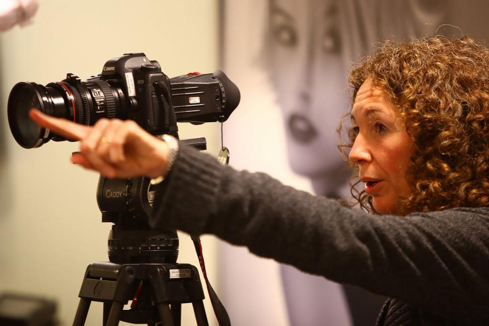 A white woman with curly hair stands behind a film camera and points.