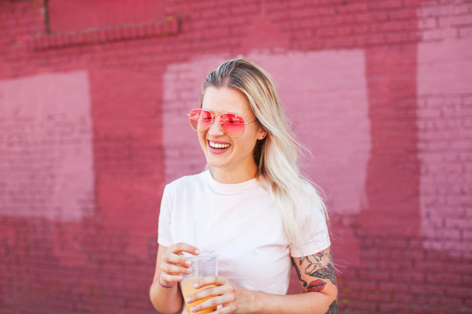 Young blonde woman with tattoos wearing pink sunglasses holding a plastic cup of juice laughing in front of pink wall