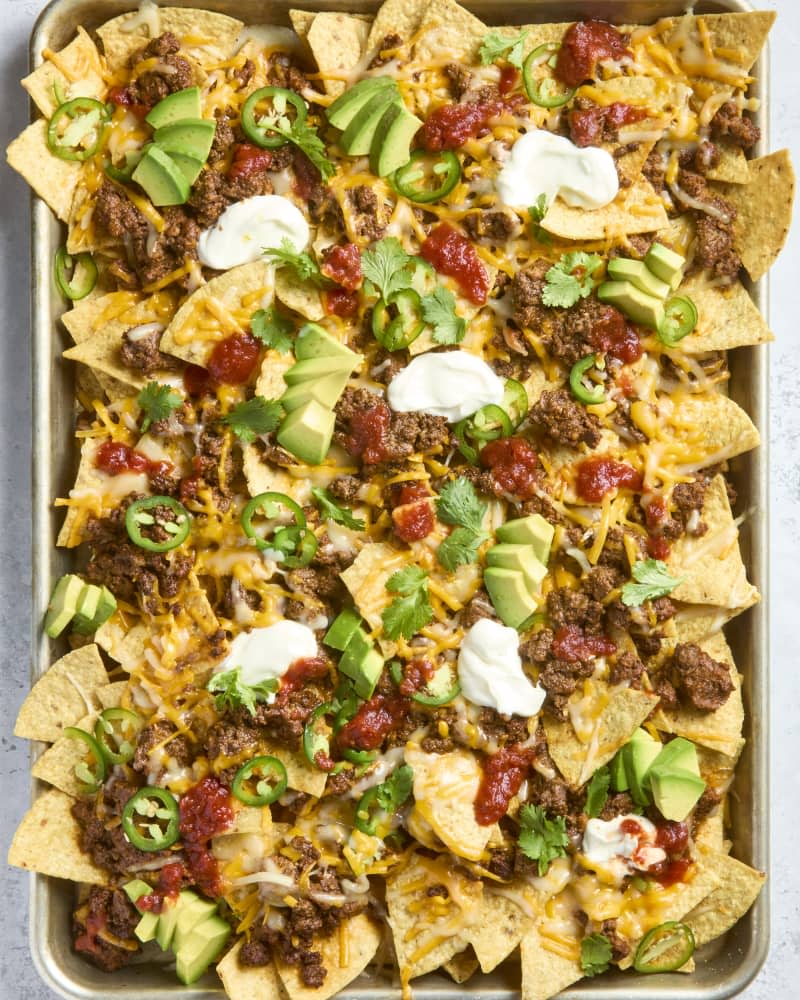 overhead shot of nachos in a sheet pan,  topped with cilantro, avocado, sour cream and salsa.
