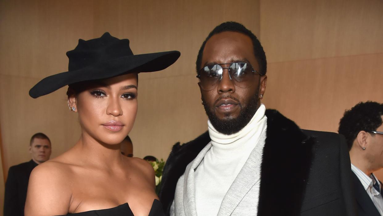 cassie and sean diddy combs stand together and pose for a photo, she wears a black hat and black strapless dress, he wears a black jacket over a gray suit and white turtleneck