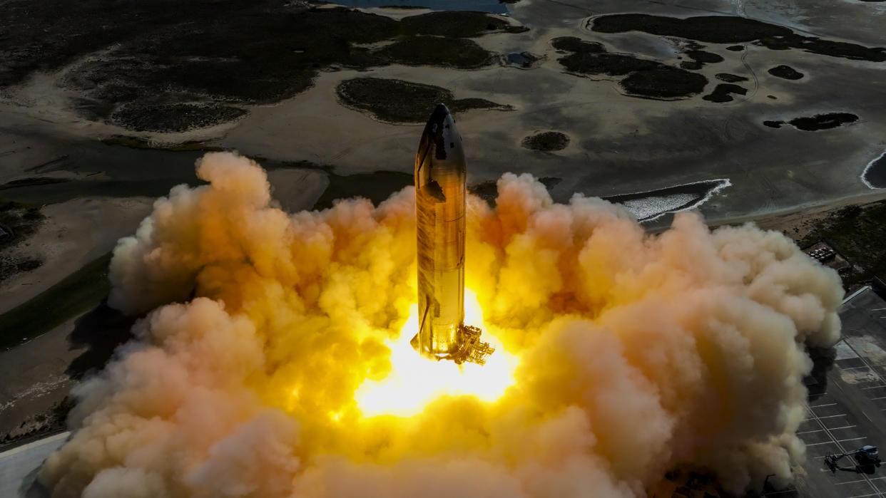  A large silver rocket on a launch pad fires its engines, creating a massive plume of fire and smoke. 