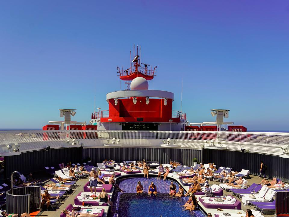 A pool on a cruise ship with people inside it and on pool chairs around it.