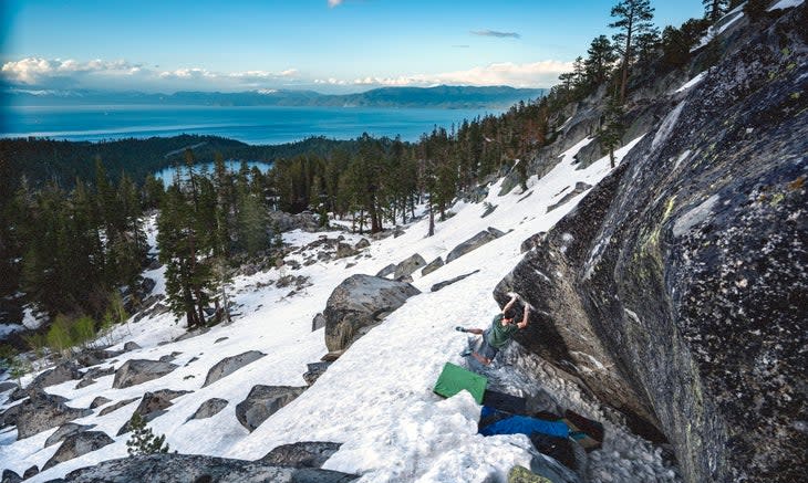 A zoomed out shot showing Jimmy Webb climbing a steep boulder above a snowy talus field with Lake Tahoe in the background.