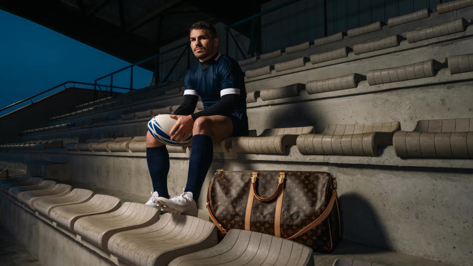 Rugby player Antoine Dupont sits in a stadium next to a Louis Vuitton monogram Keepall bag.
