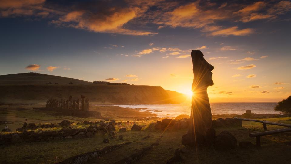 one moai statue  in the foreground has beams of sunlight radiating from behind and there is a row of moai statues in the background.