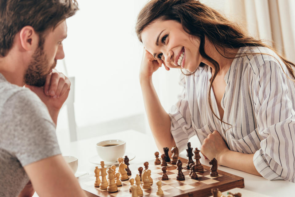 Man and woman play chess at a table, smiling at each other in a flirtatious manner