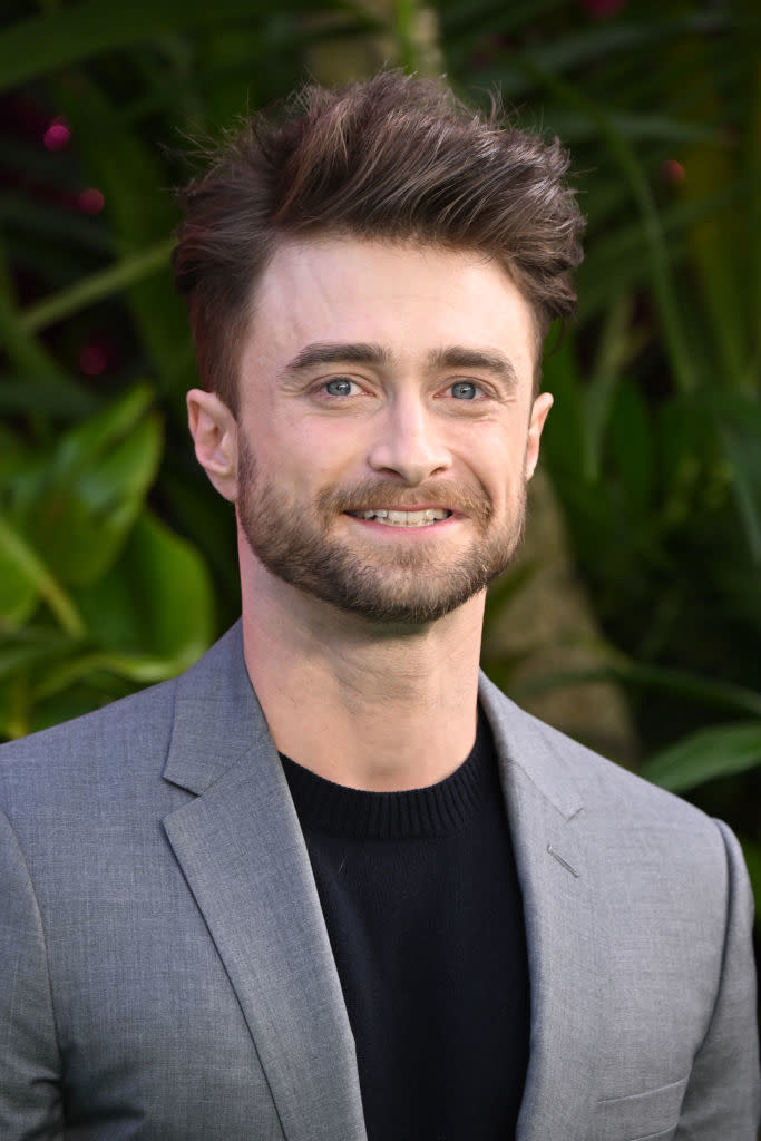 Daniel Radcliffe smiles, wearing a gray suit jacket over a black top, with a lush greenery background