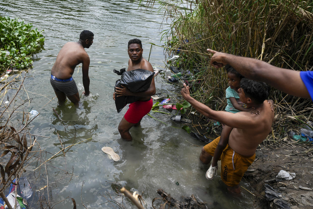 Two migrants, wearing only their underpants, and one carrying a black garbage bag, head into the river while two other migrants, one holding a young girl in his arms, point to the other side.