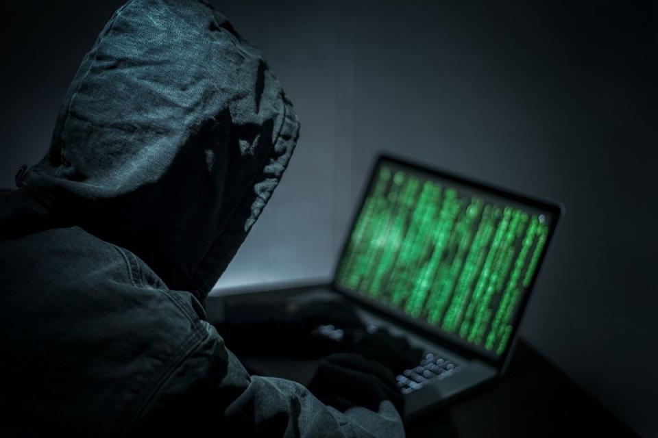 Hacker sits at laptop wearing a black hood.