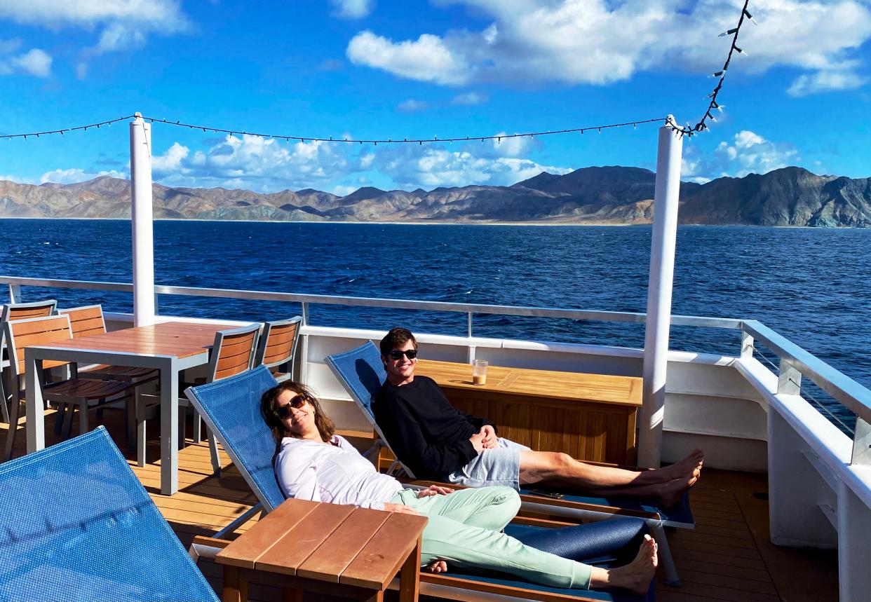 A man and woman lounge on the deck of a ship. Blue sky, mountains, and ocean water can be seen in the background.