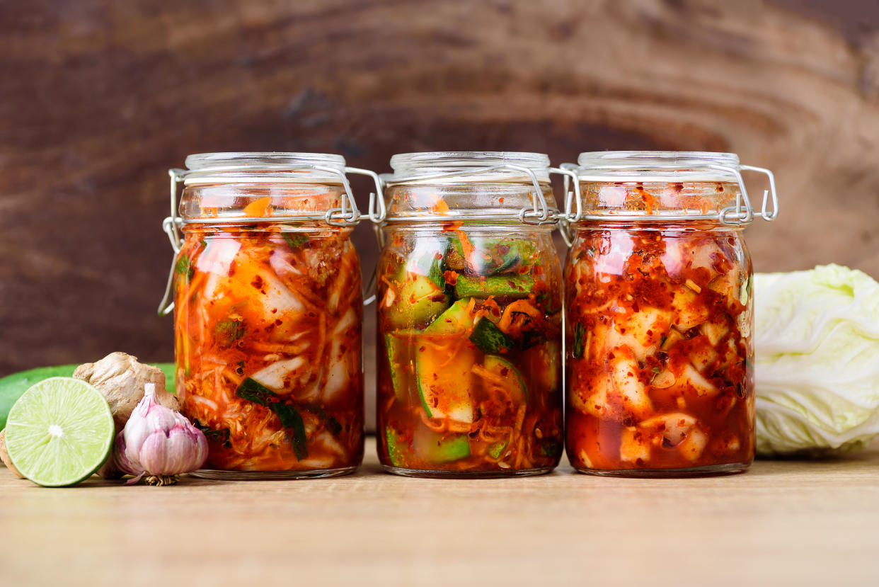 Three glass jars filled with kimchi on a table, next to half a lime, bulbs of garlic and a head of cabbage.