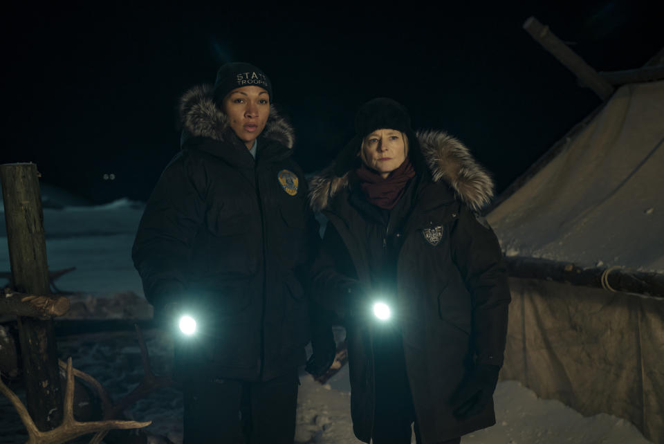 Two women in thick winter coats and hats holding flashlights while out in the snow at night; still from 