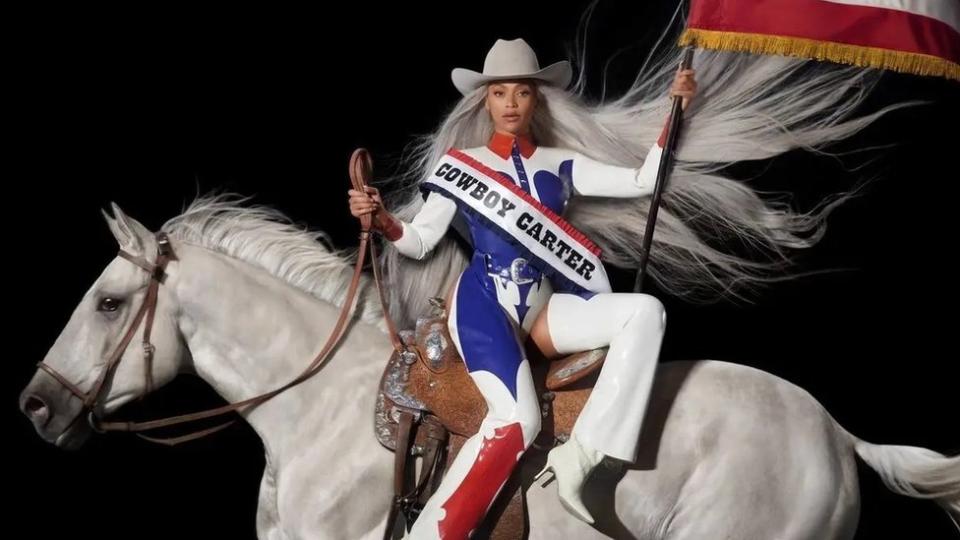 Cover of Beyoncé's album, Cowboy Carter.  Beyoncé is a 42-year-old black woman.  She wears a patent red, white and blue cowboy-themed outfit including a cowboy hat, leggings and button-down shirt.  She is shown sitting side-saddle on a white horse, her long silver hair flowing behind her.  In her right hand she holds the reins of the horse and in her left she waves the American flag.