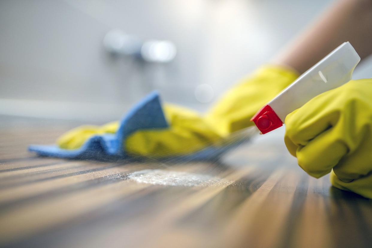 closeup of yellow gloved hands cleaning wooden table with cloth and cleaner, background out of focus