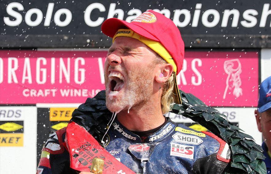 Davy Todd celebrating winning at the Southern 100, he has a yellow and a pink cap on his head, and a green garland has been draped over around his neck. He's holding a bottle of champagne with liquid from it being sprayed into his open mouth and running down his face. His mouth is open and he’s wearing blue, red and white leathers. There is a black banner with the words Solo Championships behind him and other sponsorship banners in pink, yellow and white.