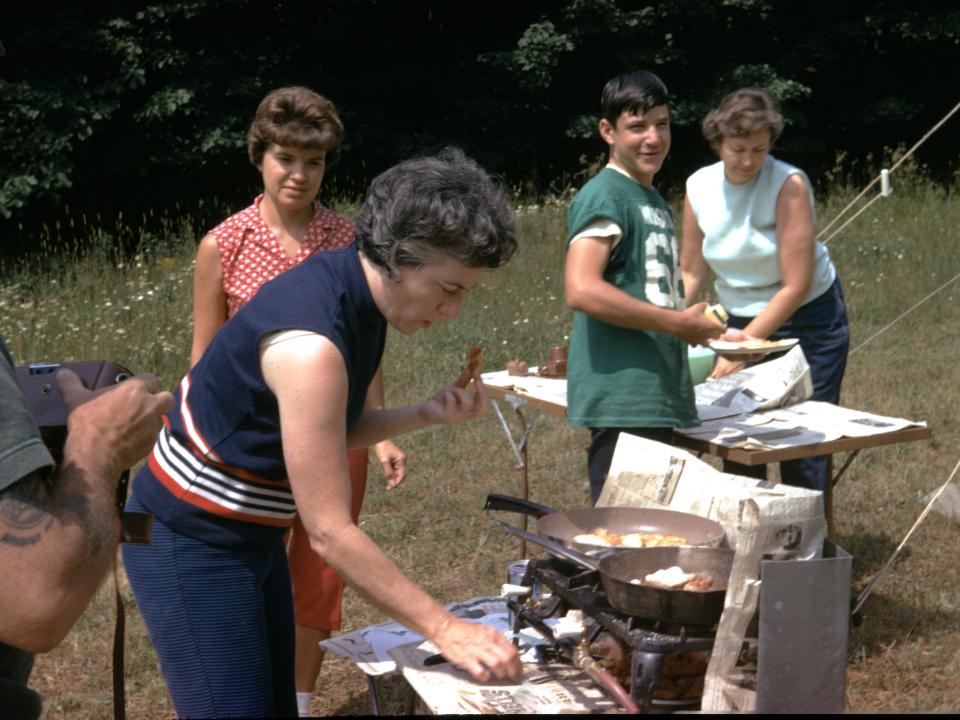 Family gathers around a camp stove at a campsite in Wyoming, United States, with mother preparing food on the stove, outdoors, 1965