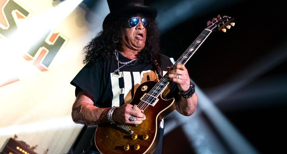  Slash plays a Gibson Les Paul onstage, against a backdrop of white stage lights. 