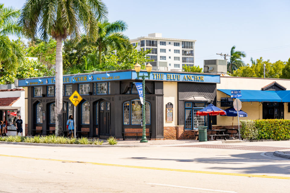 Street view of "The Blue Anchor" restaurant and bar with palm trees, pedestrians, and a nearby tall building