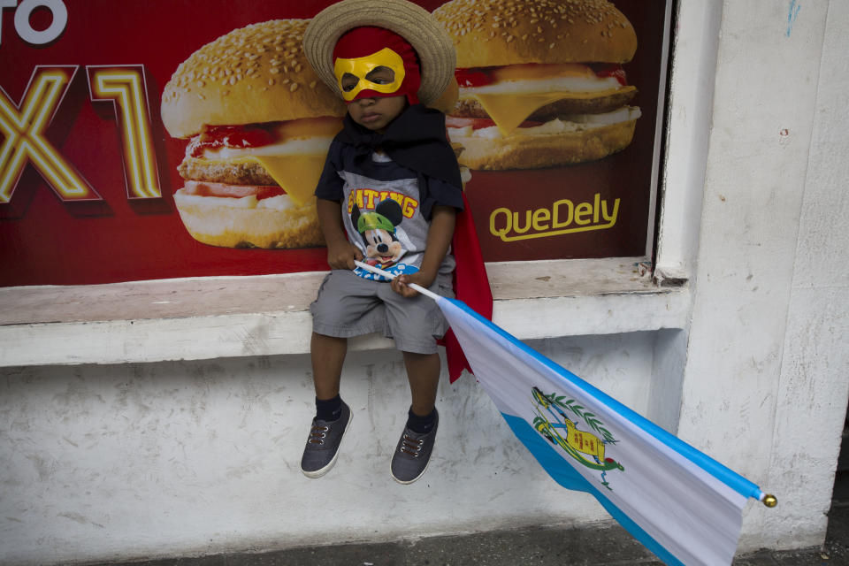 A boy in costume clutching a Guatemalan flag sits outside a local fast food restaurant as he waits with his father for a march to start against Guatemalan President Jimmy Morales and corruption in Guatemala City, Thursday, Sept. 20, 2018. Thousands marched to protest Morales' decision to end the work of a U.N. anti-corruption commission that has helped lead high-profile graft probes targeting dozens of powerful people, including one involving Morales. (AP Photo/Moises Castillo)