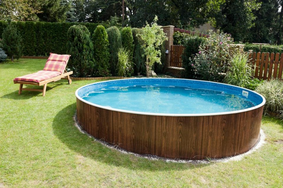 Above-ground pool with a wooden surround on a sloping lawn featuring several privacy plants