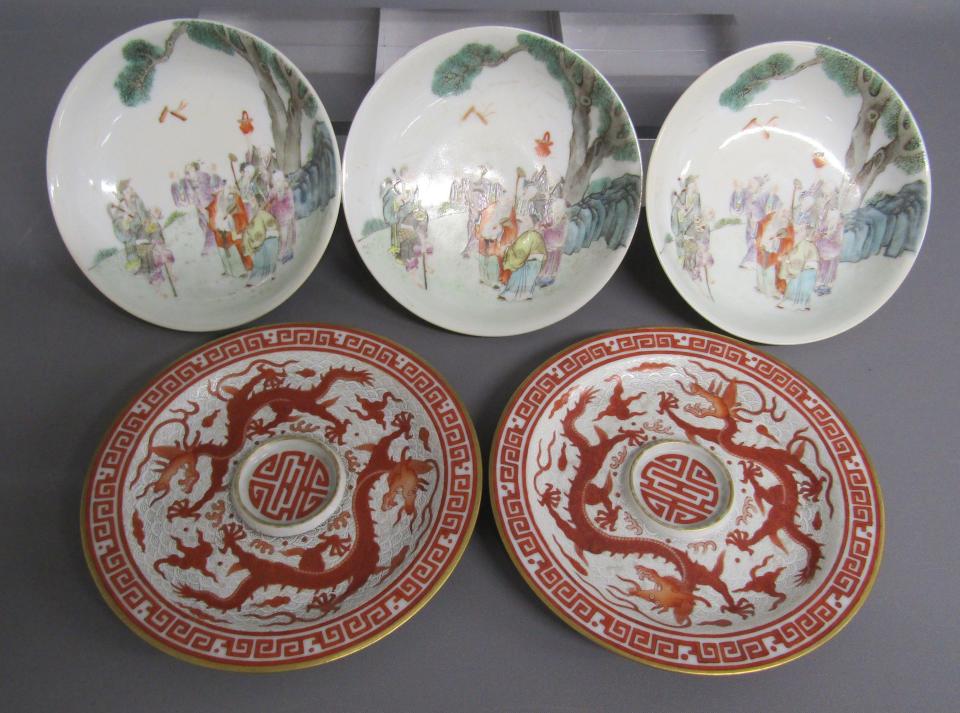 A set of five Chinese ceramic saucers, three white with a delicate multicolored depiction of a group of people in a countryside scene, and two white with a bold red pattern and dragon design.