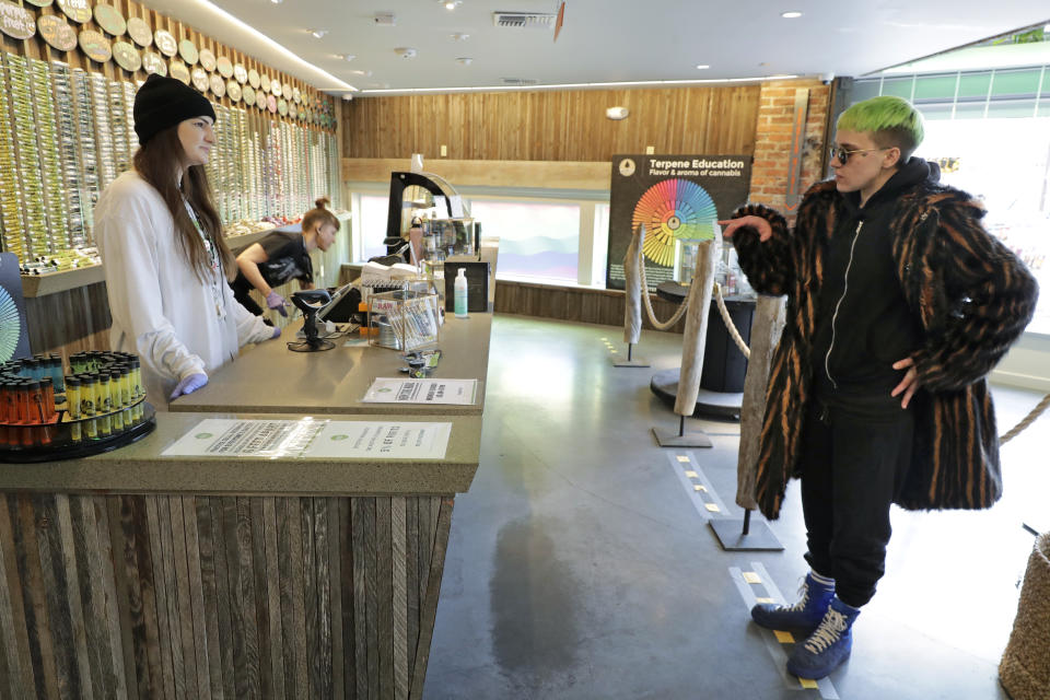 In this photo taken March 24, 2020, Ray Ray, right, talks with budtender Peyton Beech, left, as he stands on a line taped on the floor to designate a social distancing separation due to the new coronavirus while shopping at The Reef Capitol Hill, a marijuana store in Seattle. Earlier in the week, Washington Gov. Jay Inslee ordered nonessential businesses to close and the state's more than 7 million residents to stay home in order to slow the spread of the new coronavirus. In Washington and several other states where marijuana is legal, pot shops and workers in the market's supply chain were deemed essential and allowed to remain open. (AP Photo/Ted S. Warren)