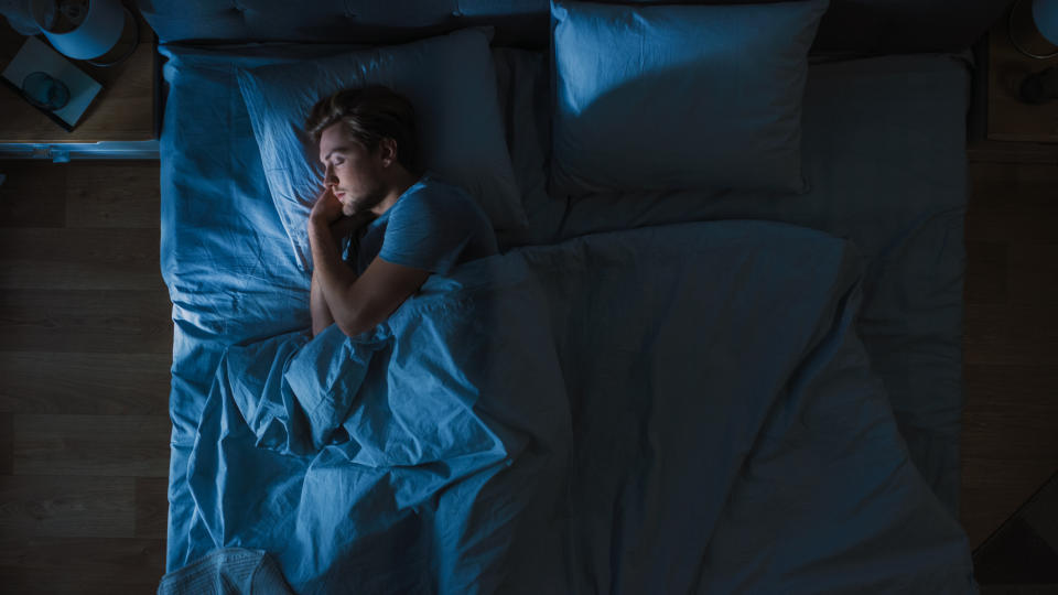 Top View of Handsome Young Man Sleeping Cozily on a Bed in His Bedroom at Night. Blue Nightly Colors with Cold Weak Lamppost Light Shining Through the Window.