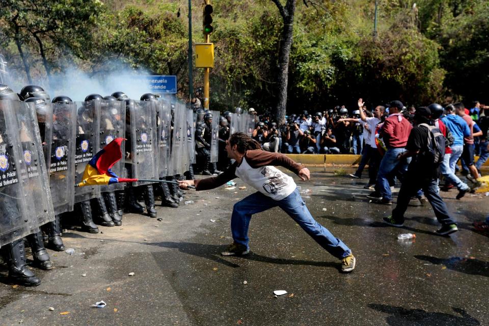 A demonstrator uses a Venezuelan flag to strike at a line of Bolivarian National Police officers in riot gear, during clashes at a anti-government protest in Caracas, Venezuela, Wednesday, March 12, 2014. A university student has died and a number of others, including three National Guardsmen, have been wounded after being shot by unknown assailants in two separate incidents in the central Venezuelan city of Valencia. (AP Photo/Fernando Llano)