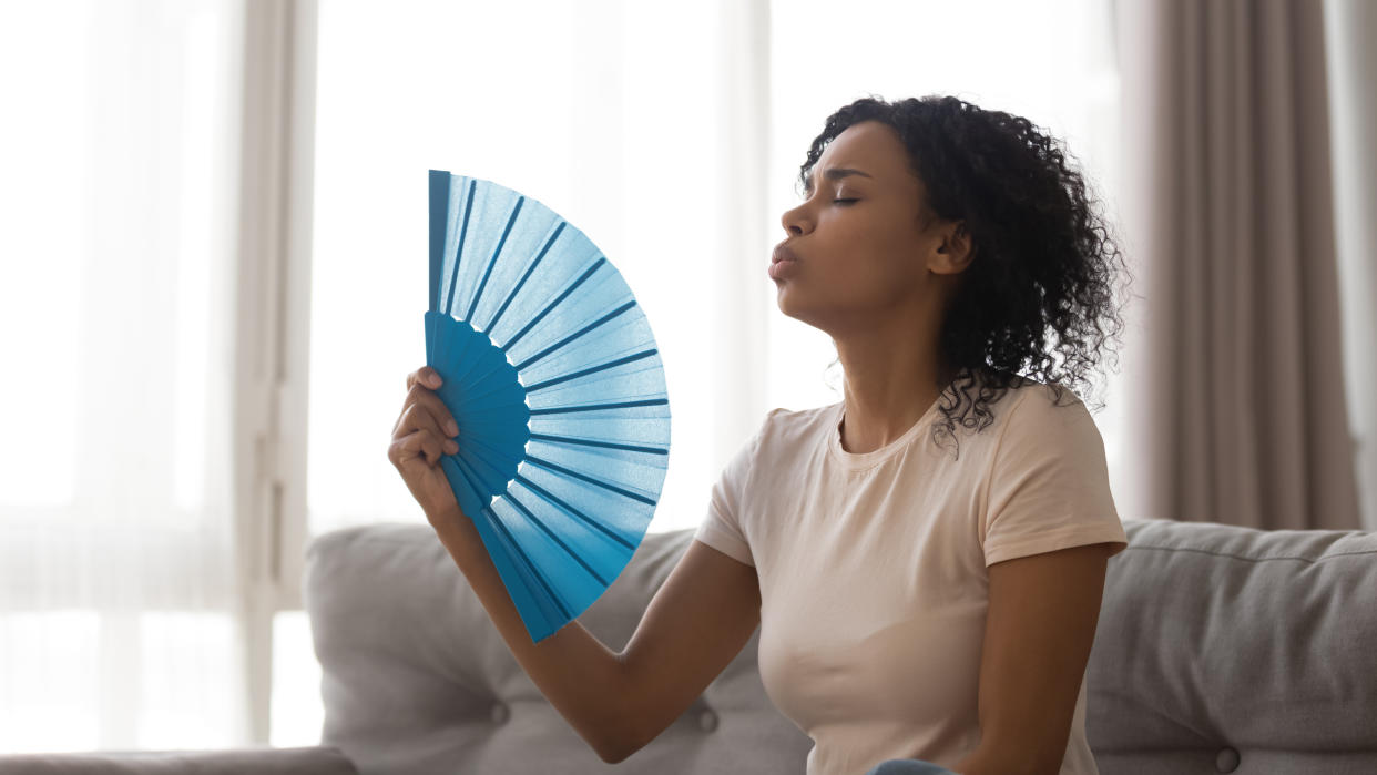 African woman sitting on couch in living room alone cools herself with blue handheld fan waved back-and-forth in hot weather at home without air conditioning, airflow relief heat helps feels refreshed