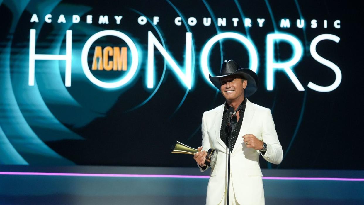 tim mcgraw smiles and stands behind a microphone while on a stage, he wears a white suit with a black collared shirt with white polka dots and a black cowboy hat, he holds a gold award in one hand, behind him is a background that says academy of country music honors