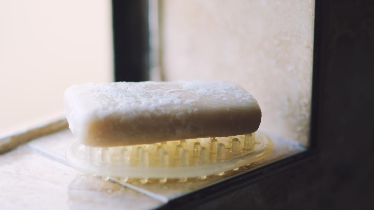 Bar of soap textured with fresh suds sits on a soap dish on a shelf at the shower window of a dark bathroom.