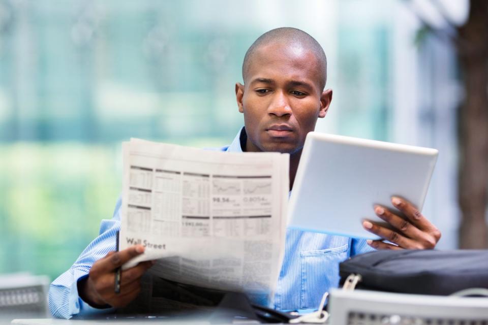 A seated person reading a financial newspaper while holding a tablet in their left hand.