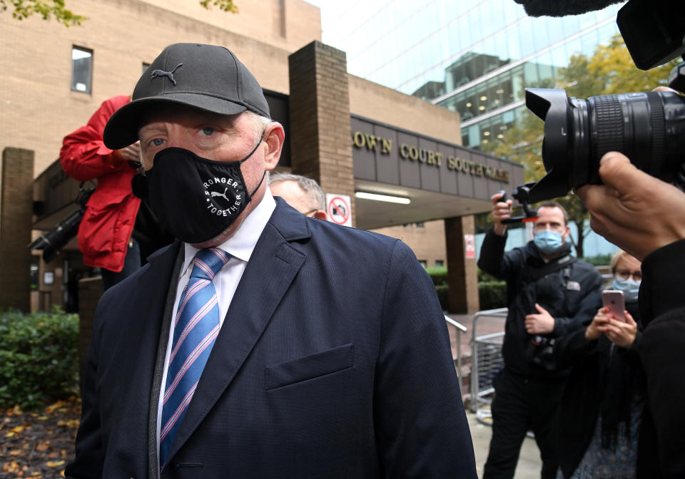 Boris Becker, in a mask, suit and baseball hat, outside of a town court building with cameras in his face. 