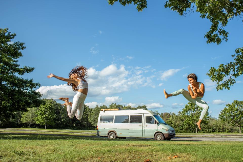 Nat and Abi jump in the air in front fo their van with blue skies behind them.