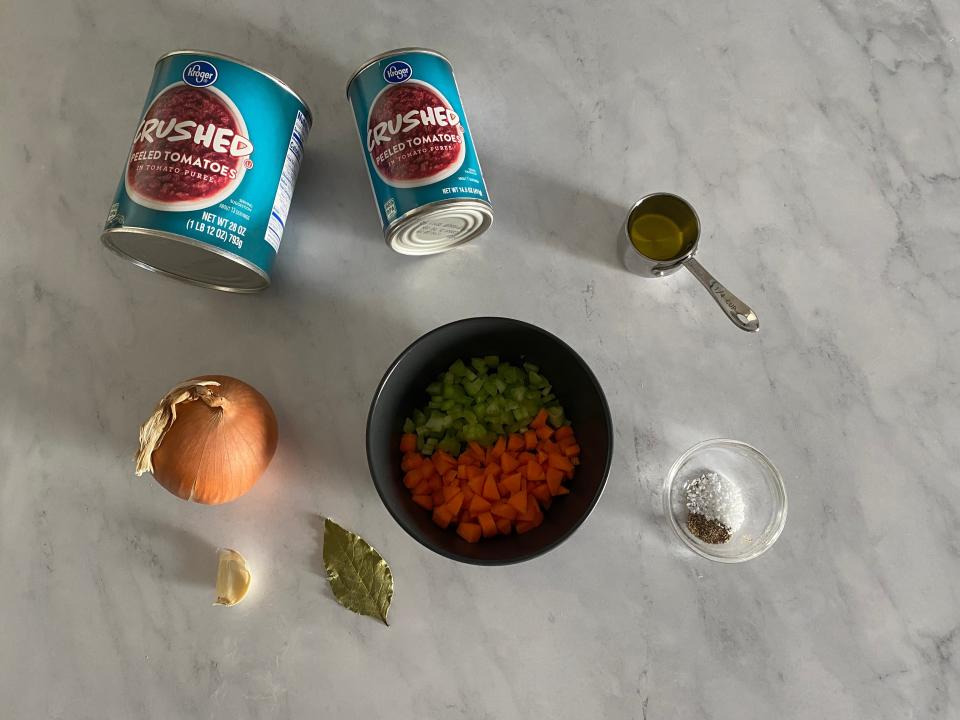 Two cans of crushed tomatoes, bowl of diced carrots and celery, inion, a garlic clove, a bay leaf, and spices on a table