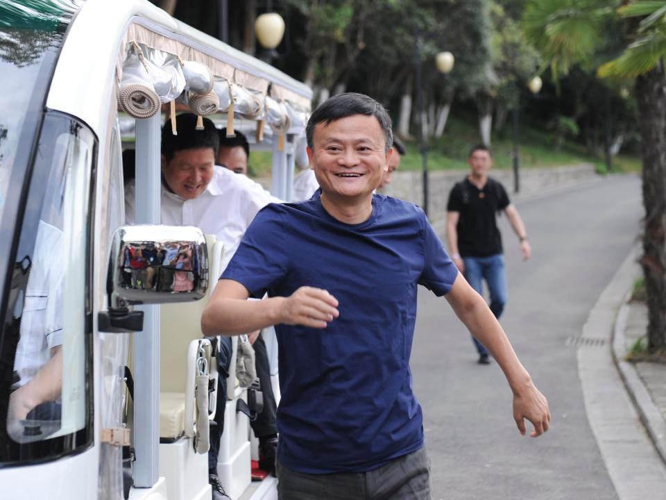 Jack Ma running down a road alongside a white car while wearing a blue T-shirt and smiling.