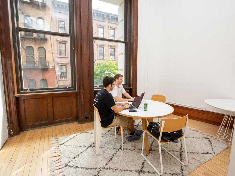 Two people working at a round table on a patterned rug near large windows.