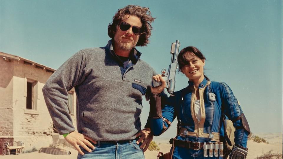 A young woman in a futuristic blue jumpsuit smiles as she wields an oversized, science-fiction style silver blaster gun. She's standing next to a man in a grey fleece, hands on his hips, who wears sunglasses as his mid-length hair is ruffled by the wind. They appear to be in a desert setting, with blue sky behind them and bright sun illuminating the scene