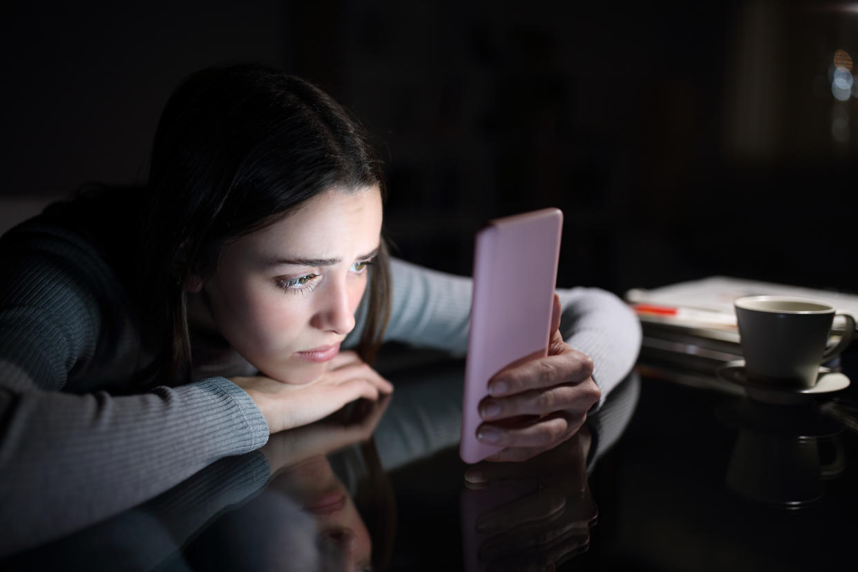 A sad-looking woman rests her chin on her hand, which is placed on a table, as she looks glumly at her phone with her other hand