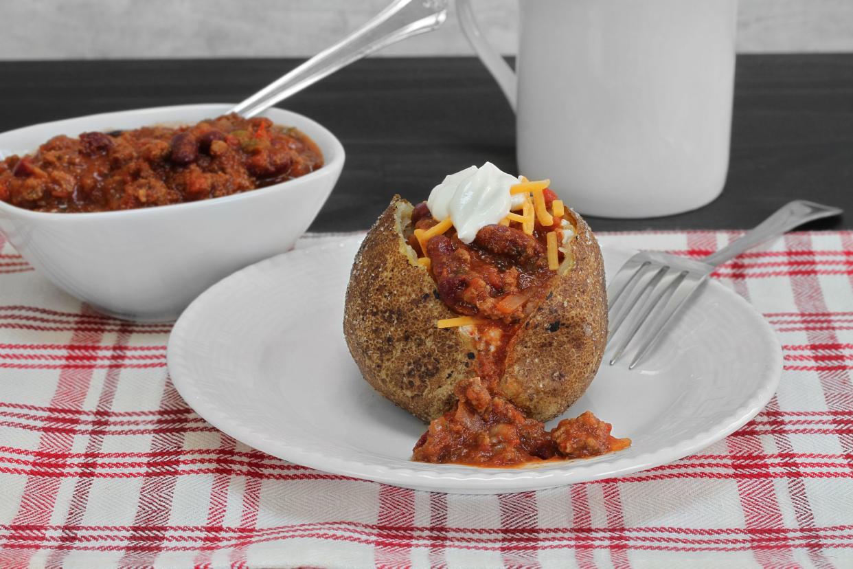 One baked potato topped with chili, cheddar cheese and sour cream. Macro, close up.  Background includes a bowl of chili.