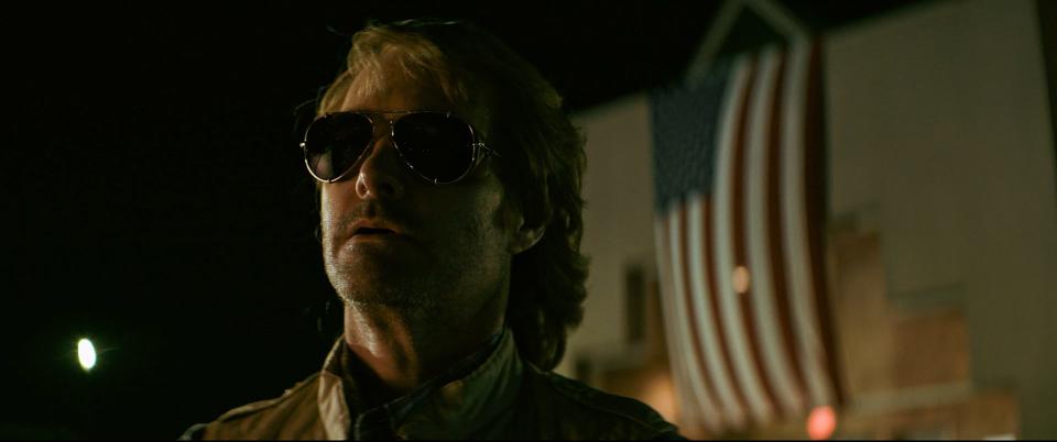 Will Forte's MacGruber with sunglasses stands in front of an American flag
