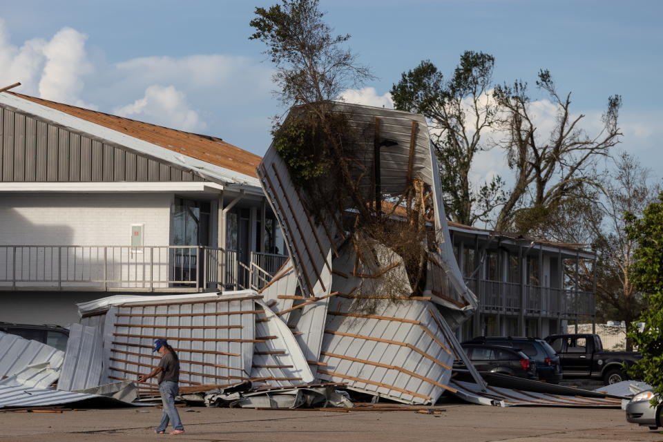 A person walks debris including a mangled section of roof that rests in a parking lot.