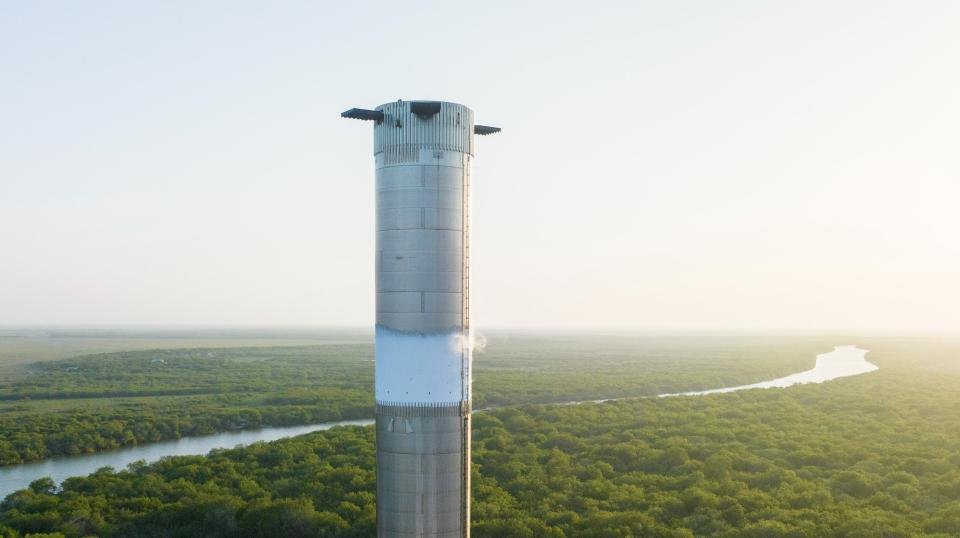 A silver SpaceX Starship Super Heavy Booster prototype sits against a background of green trees and a meandering river.