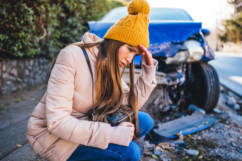 Frustrated young woman crouches next to wrecked car after a car accident.