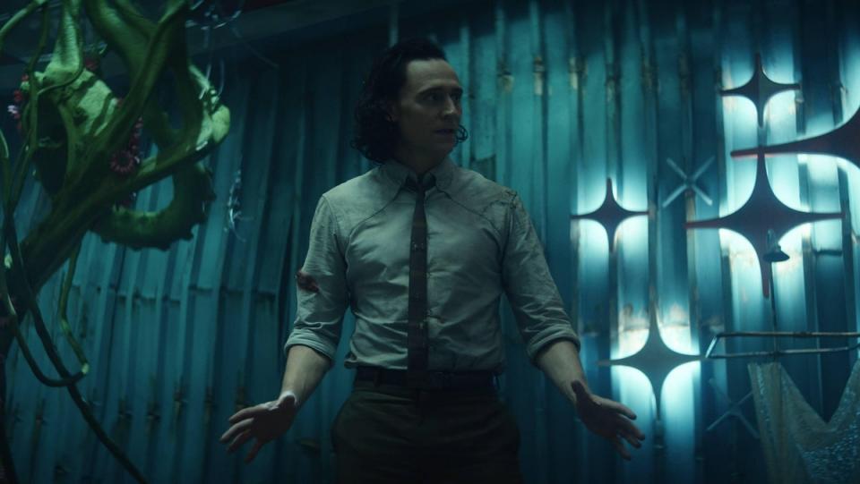 Loki stands looking very concerned, arms prepared for action. He wears his button-up TVA shirt and dark skinny tie. He stands in front of the sparkly bowling alley wall.