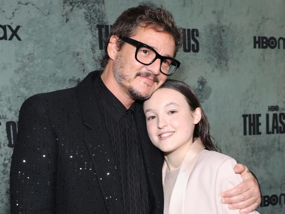 pedro pascal and bella ramsey as the last of us premiere. pascal is holding ramsey to his chest with his left arm, resting his chin on top of their head. ramsey is smiling widely while wearing a blush pink suit and their hair combed back from their face, while pascal is in a shimmering black suit and black thick framed glasses