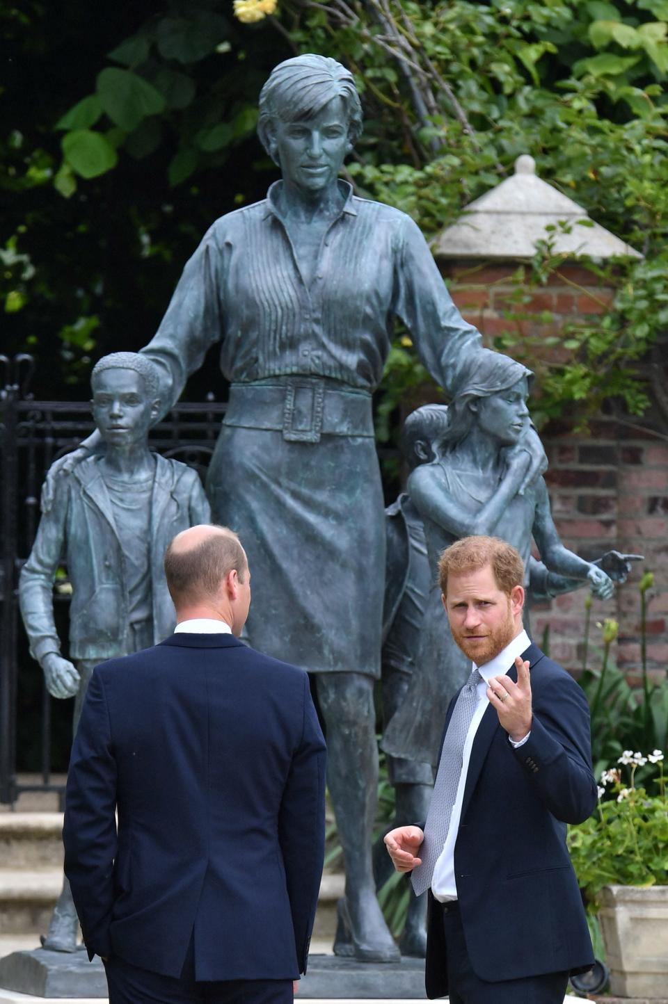 Harry speaking to William in front of a blue statue of Princess Diana.